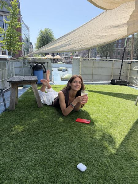 A woman is relaxing on artificial grass on a sunny day, holding a drink and smiling at the camera